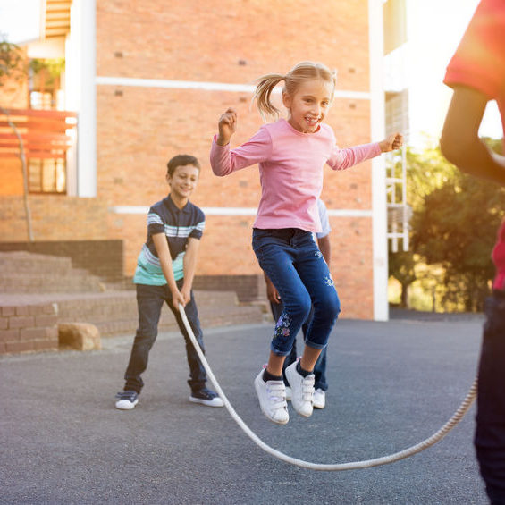 Happy elementary kids playing together with jumping rope outdoor. Children playing skipping rope jumping game and laughing outdoors. Happy cute girl jumping over skipping rope held by her friends.